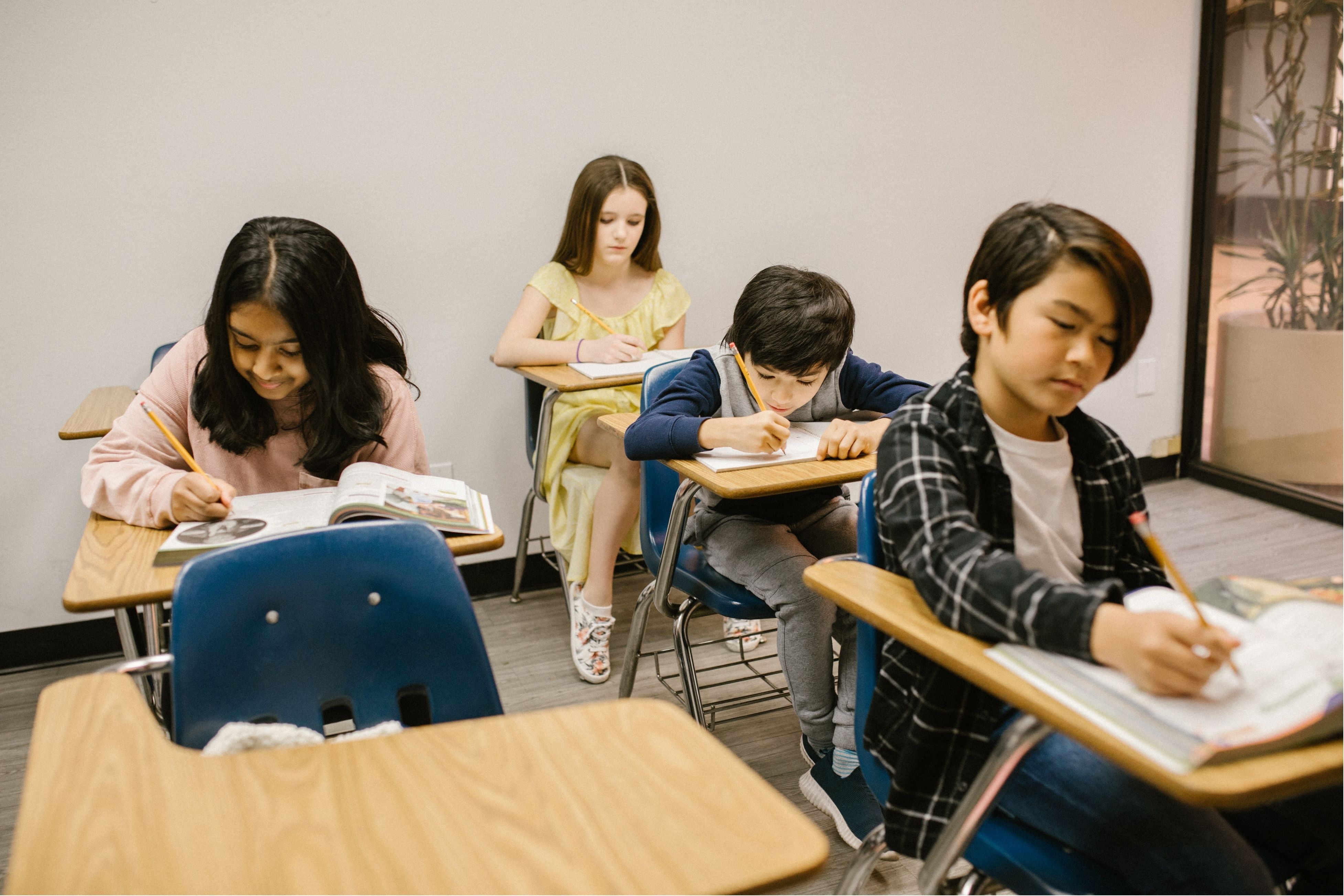 Students are sitting at individual desks in a classroom, writing in notebooks and books.