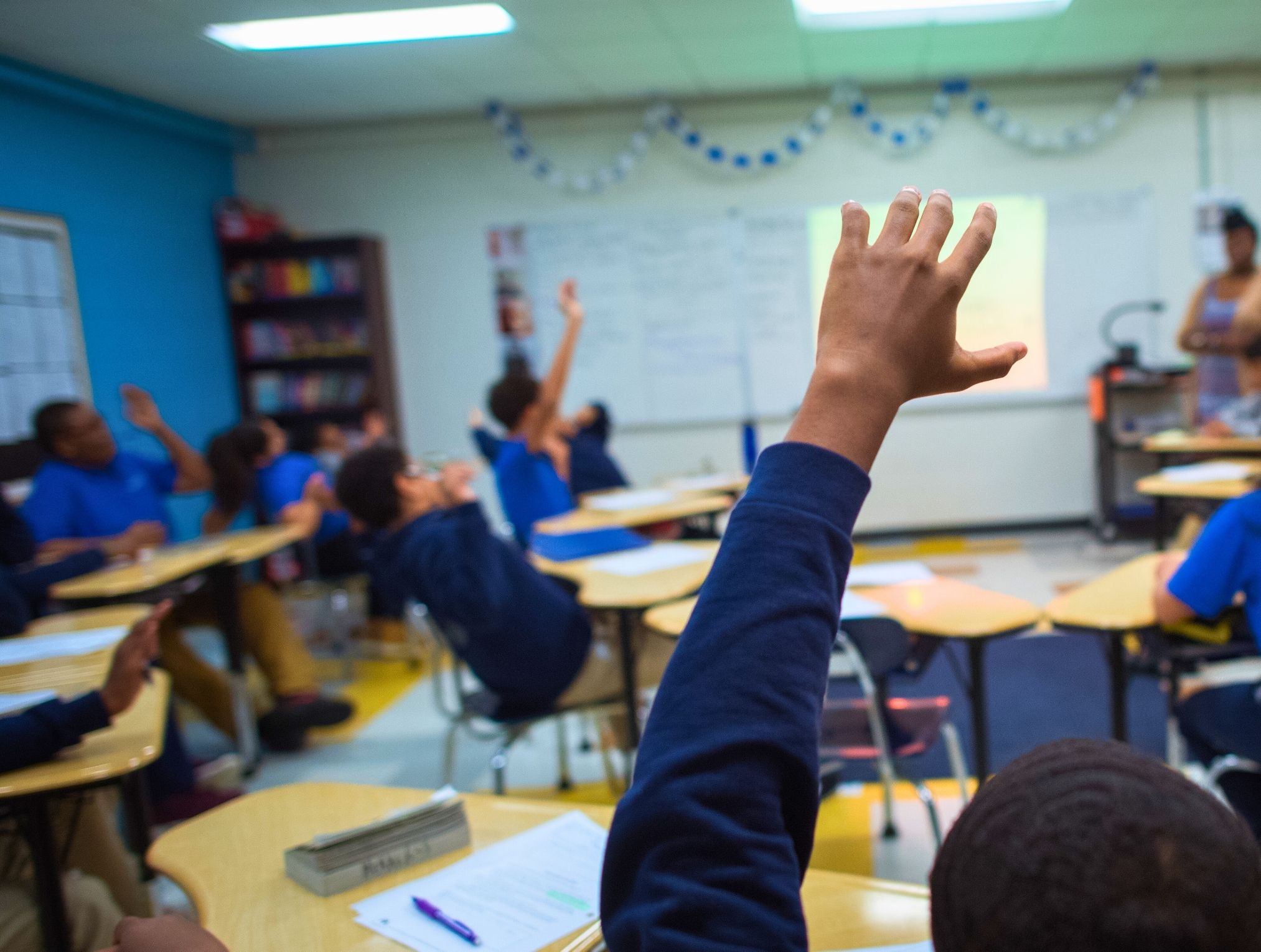 A classroom scene with students raising their hands to answer a question. Several students in blue shirts are seated at desks, with one student in the foreground raising their hand prominently.