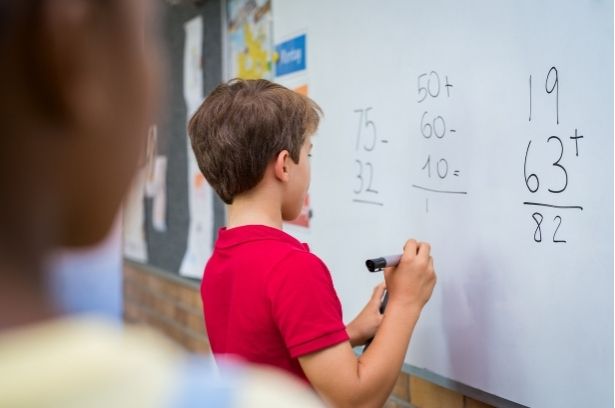 A boy in a red shirt writes math problems on a whiteboard with a marker. Math equations and numbers are visible on the board.