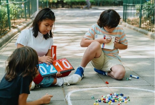 Three children sit on a paved surface, engaged in a game with numbered boards and marbles. One child holds a notebook and pen, appearing to record scores or information.