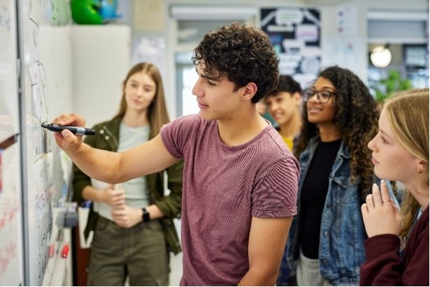 A young person with curly hair writes on a whiteboard while a group of peers watches attentively in a classroom setting.