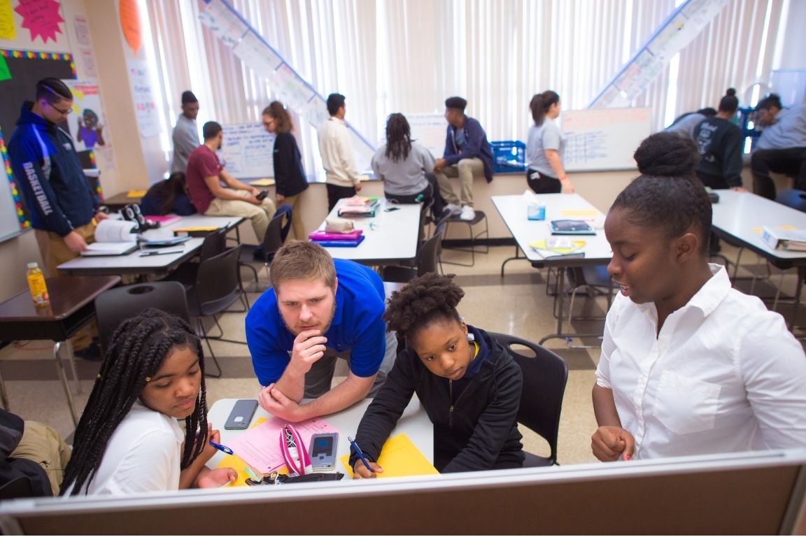 A teacher in a blue shirt leans over a table to help three students with their work in a classroom setting.