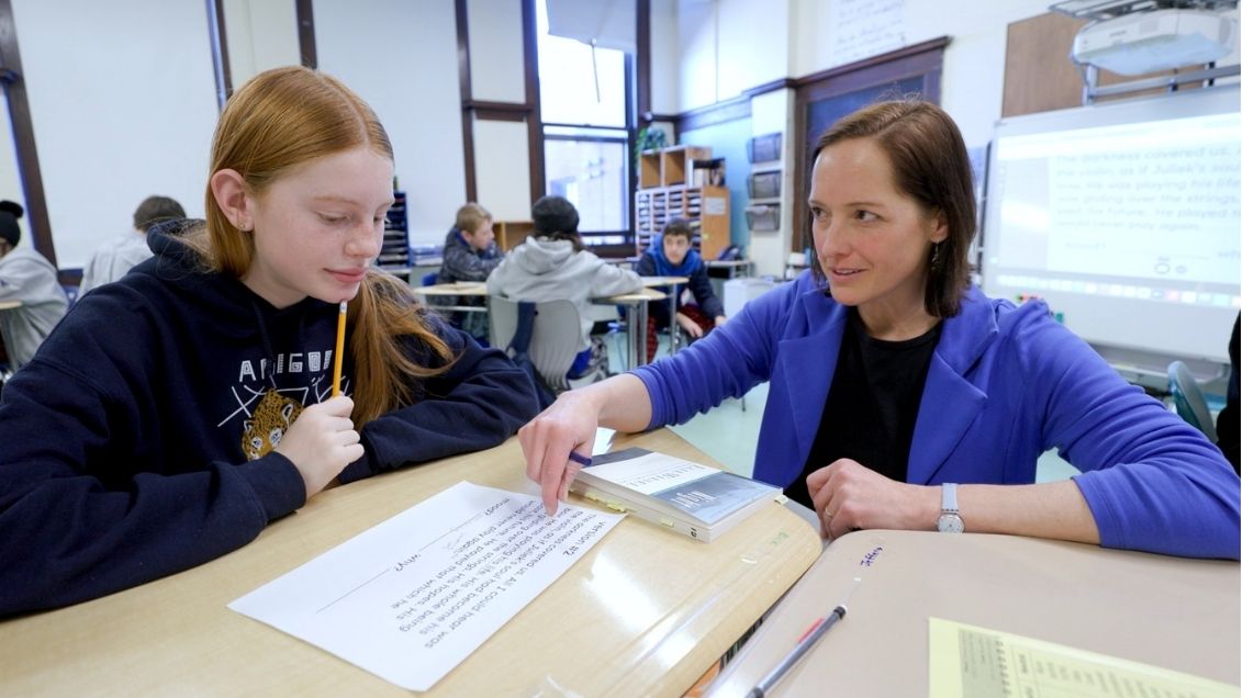 A teacher in a blue blazer points at a paper while a student with red hair holds a pencil and looks at the paper.