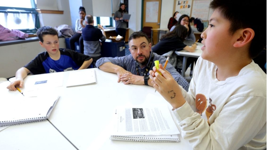 A teacher looks at students sitting around a table in a classroom, with notebooks and papers spread out.